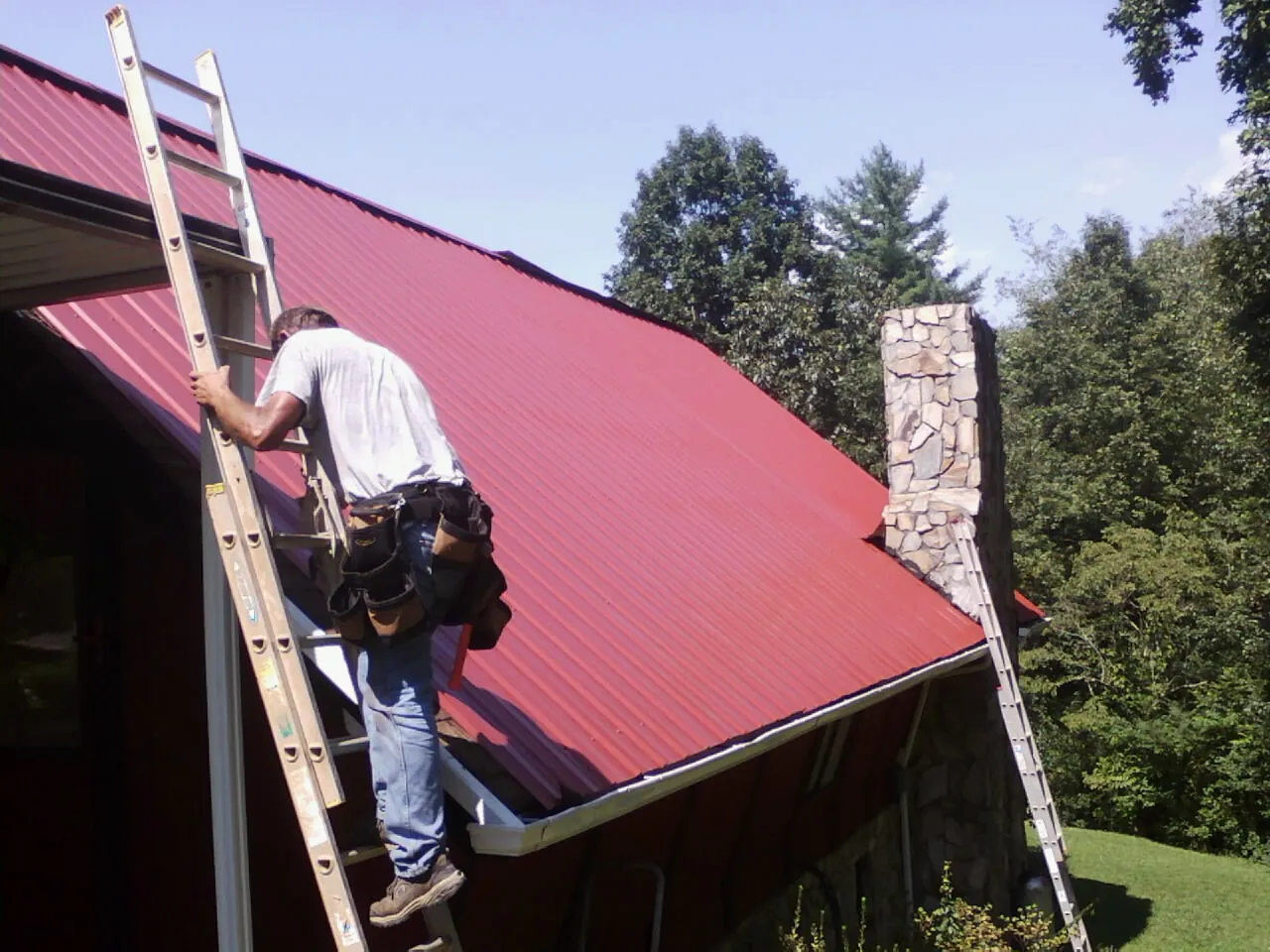 Roofer on a ladder beside a red metal roof and stone chimney — Soffit & Fascia Repair by Roofing Brothers in Lowry